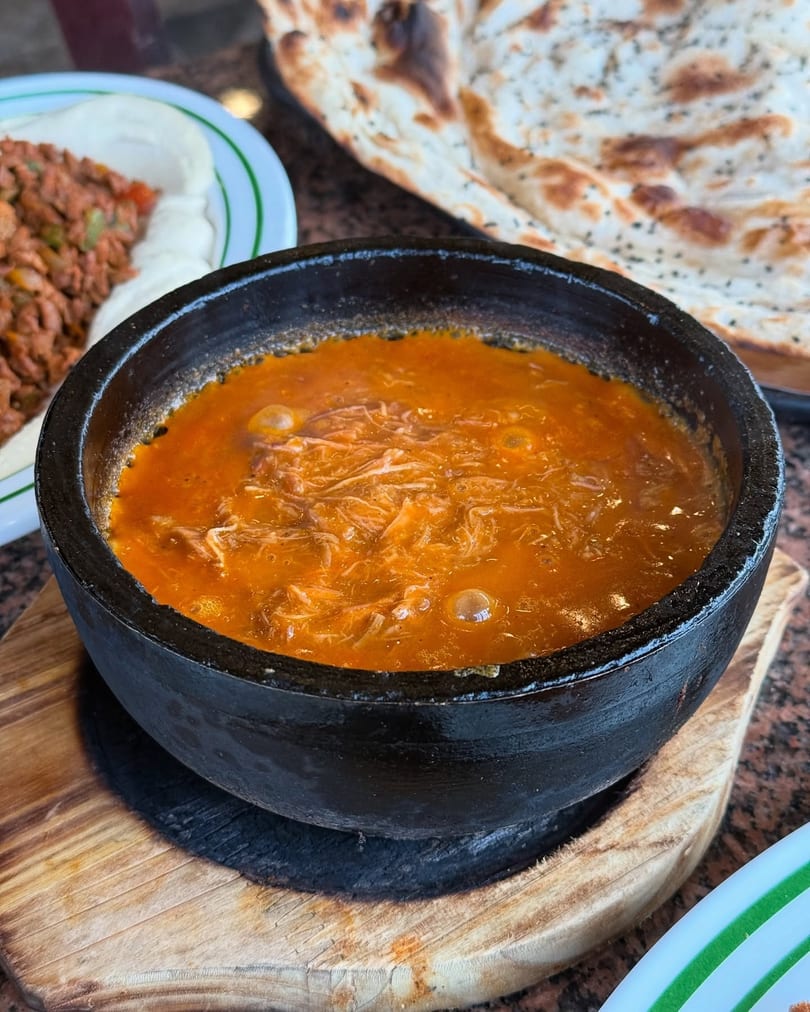 A black bowl of bubbling orange stew with shredded meat, served with flatbread and another dish.