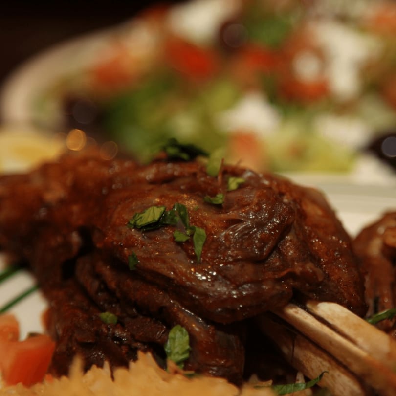 Close-up of a roasted lamb shank garnished with fresh herbs, served with rice and salad.