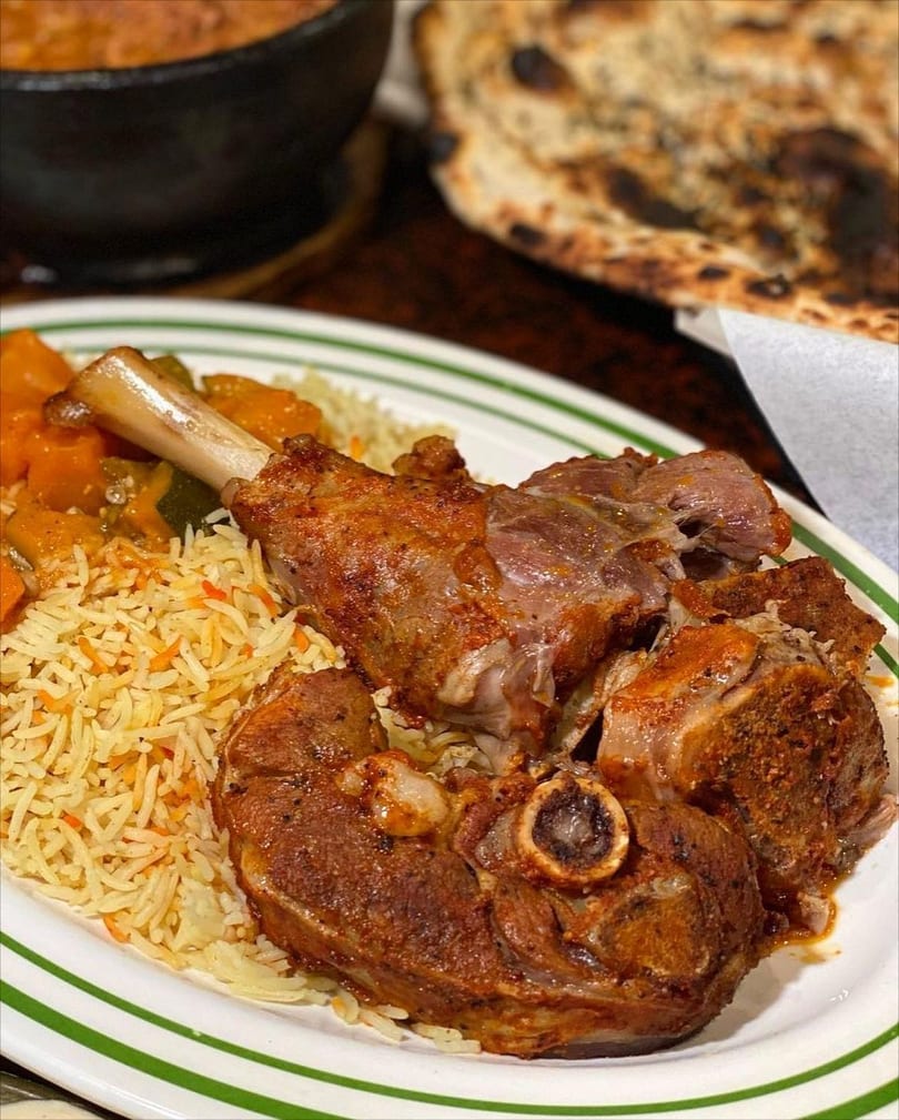 A plate of lamb shank and meat pieces with rice and vegetables, with flatbread in the background.