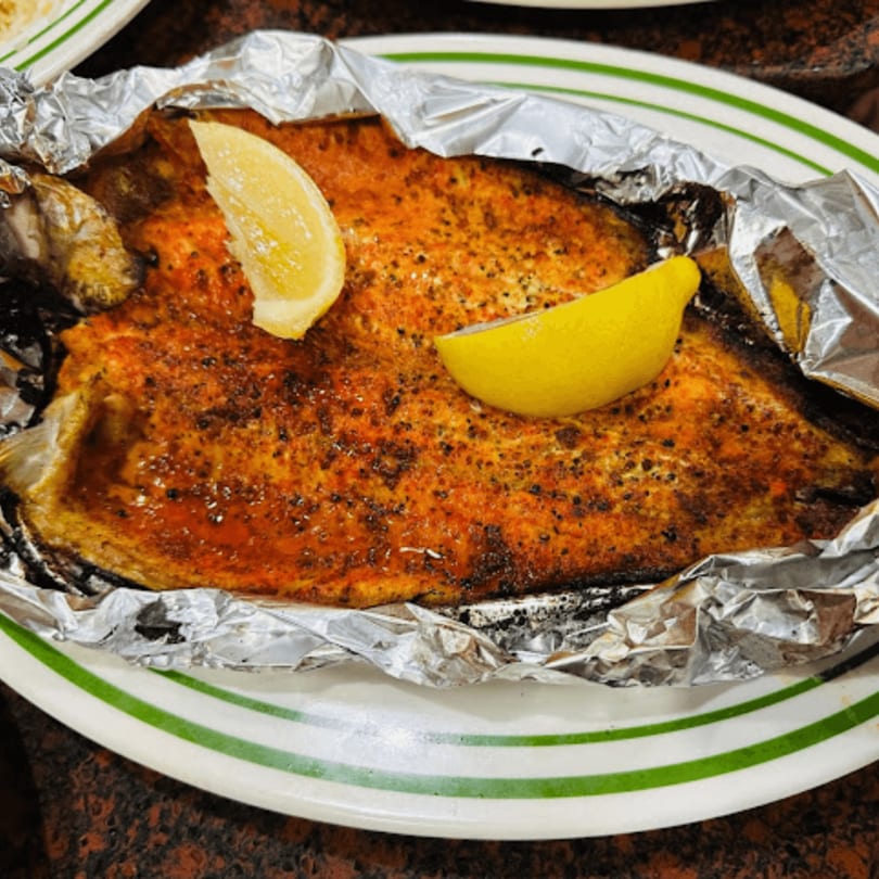 Baked fish with seasoning and two lemon wedges, served in foil on a white plate.