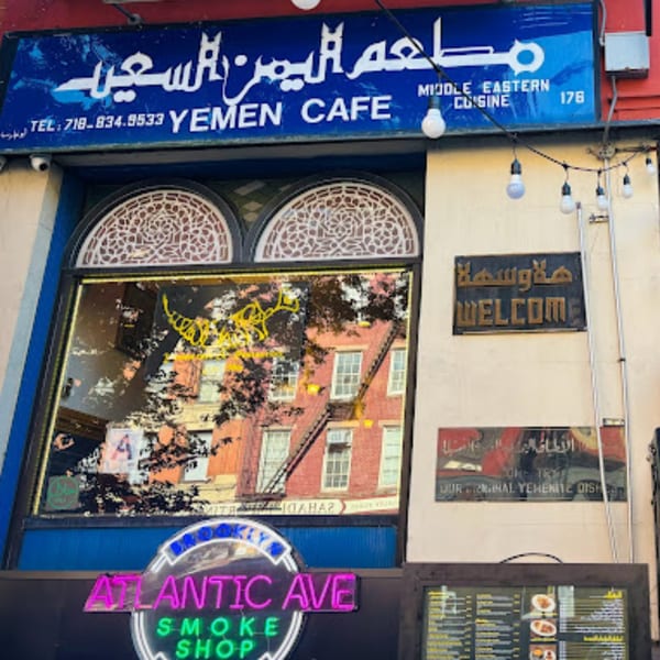 Exterior of Yemen Cafe & Restaurant with a blue sign, arched windows, and a neon "Atlantic Ave" sign.