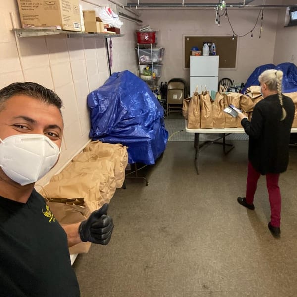 Man in a mask gives a thumbs up while a woman prepares brown paper bags of food in a storage room.