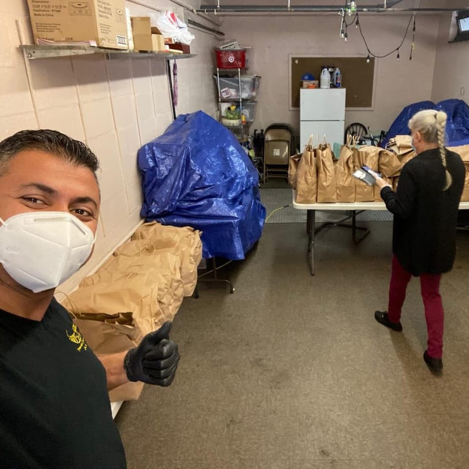 Man in a mask gives a thumbs up while a woman prepares brown paper bags of food in a storage room.