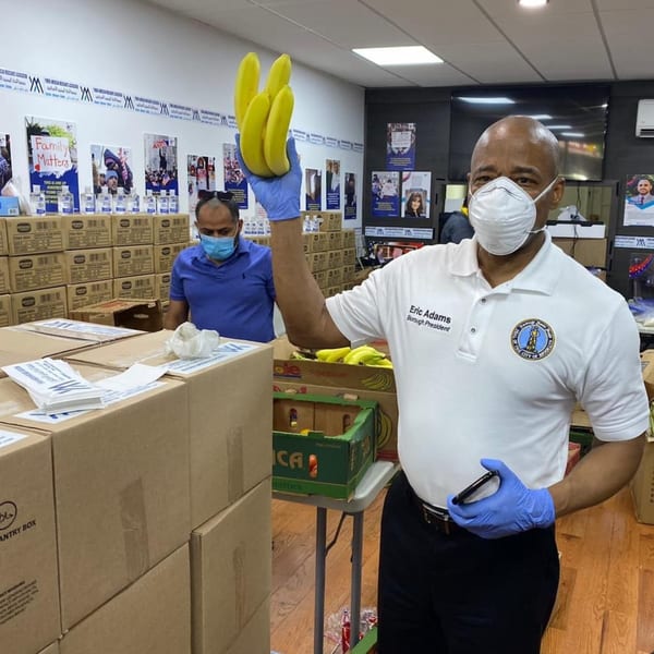 Eric Adams, Borough President, wearing a mask and gloves, holding bananas in a room with boxes.