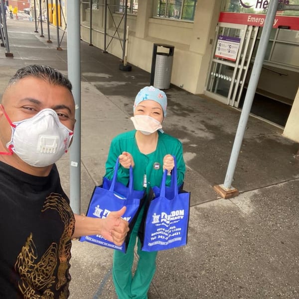 A man in a mask gives a thumbs up next to a masked healthcare worker holding two blue bags.