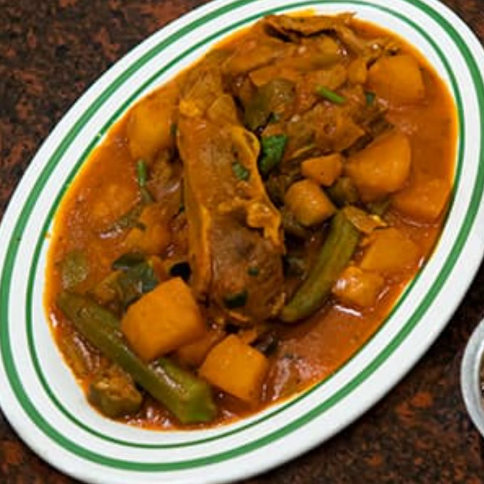 A white oval plate with green stripes holds a hearty stew of meat, potatoes, and okra in a rich orange sauce.
